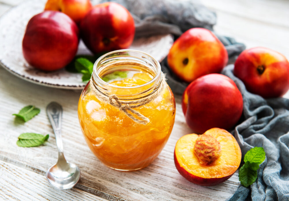 Löffel neben Glas mit Nektarinen-Marmelade in geöffnetem Gläschen steht neben frischen Nektarinen und Minze auf Tischdecke