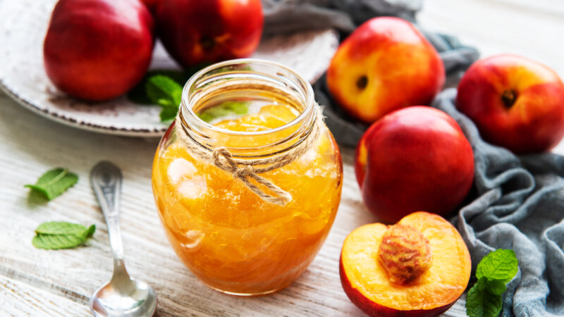 Löffel neben Glas mit Nektarinen-Marmelade in geöffnetem Gläschen steht neben frischen Nektarinen und Minze auf Tischdecke