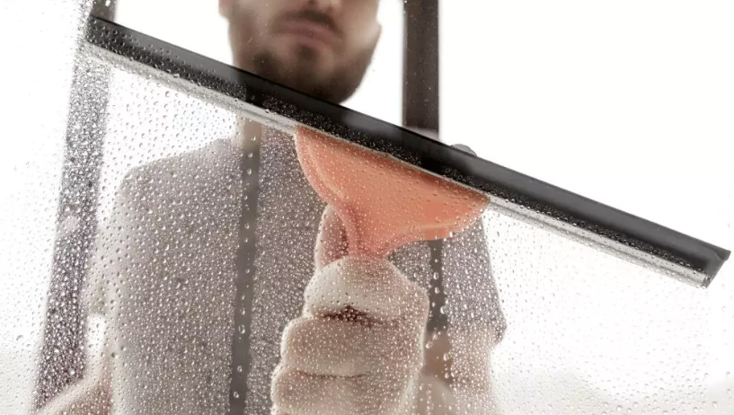 Gebäudereinigung Ender beim Fensterwischen Glasscheibe mit einem großen Fensterwischer und orangem Haltergriff zu sehen der von einem Mann gehalten wird bei dem nur der Oberkörper und der Mund sowie die Nase zu sehen sind