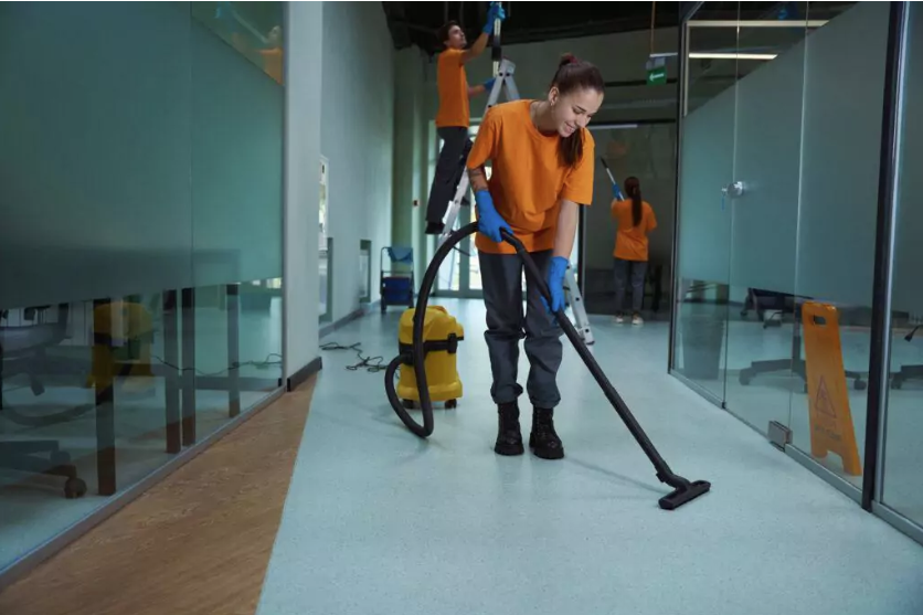 Gebäudereinigung Ender bei der Arbeit Frau mit orangem T-Shirt und schwarzer Hose hat blaue Hygienehandschuhe an und saugt den Boden