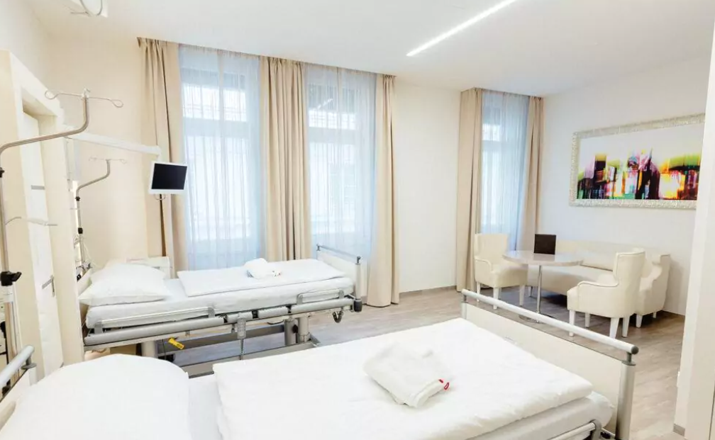 Ästhetische Chirurgie Wien Krankenzimmer mit zwei Krankenbetten in weiß. Dahinter eine Sitzgruppe mit hellen Möbel und drei großen Glasfenster mit beigen Vorhängen